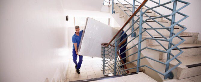 Two professional movers carry a wrapped mattress up a bright stairwell with blue railings and wide steps.