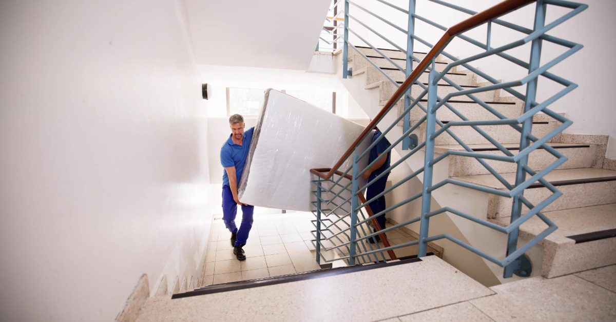 Two professional movers carry a wrapped mattress up a bright stairwell with blue railings and wide steps.
