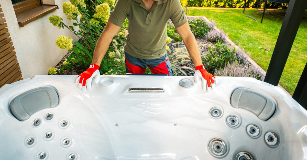A worker in red gloves steadies a white hot tub beside a garden and lawn near the back patio of a home.
