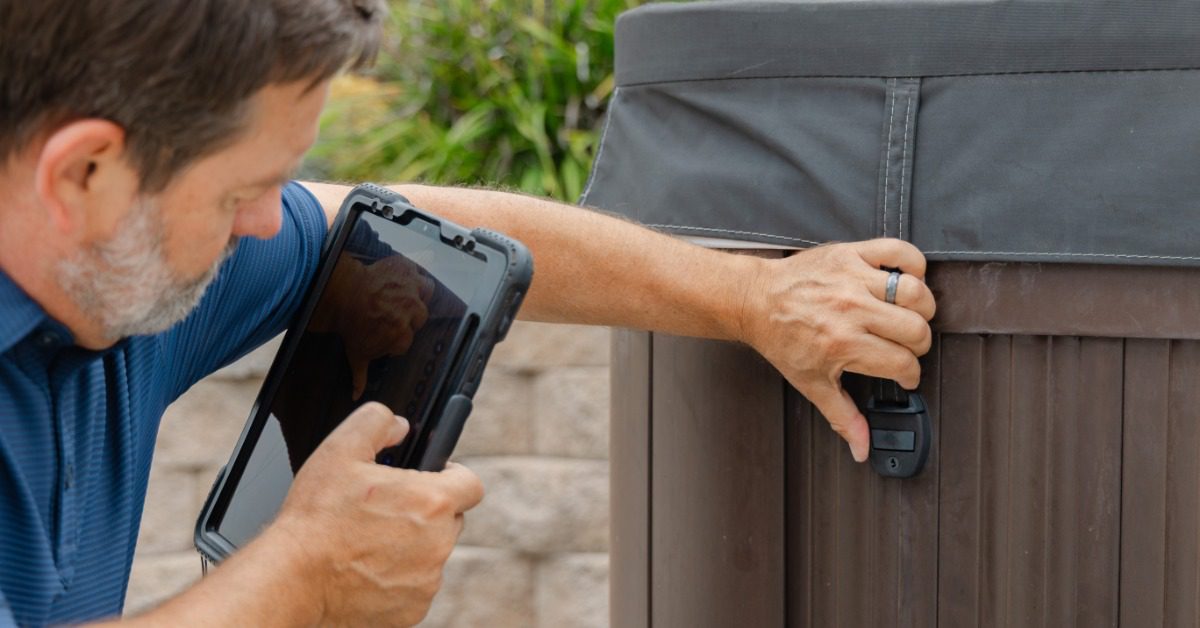 A man with a tablet photographs a hot tub cover latch on a brown spa cabinet near an outdoor stone patio.