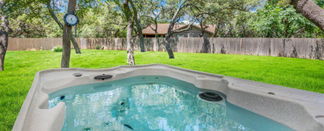 A filled hot tub sits under a large patio umbrella in a fenced backyard with trees and bright green grass.
