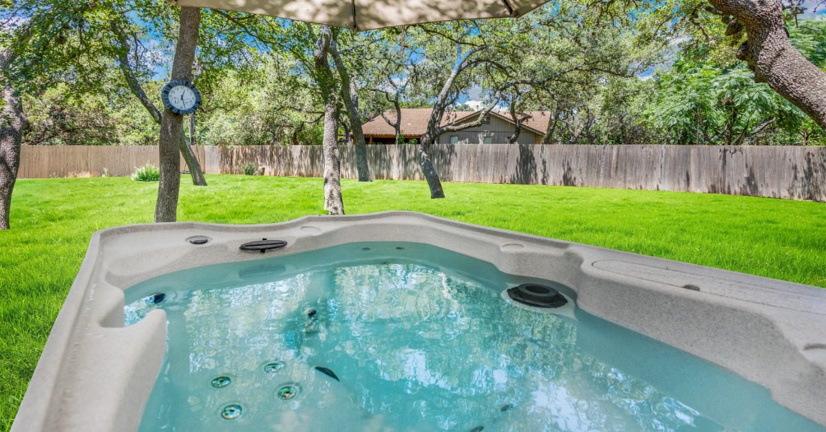 A filled hot tub sits under a large patio umbrella in a fenced backyard with trees and bright green grass.