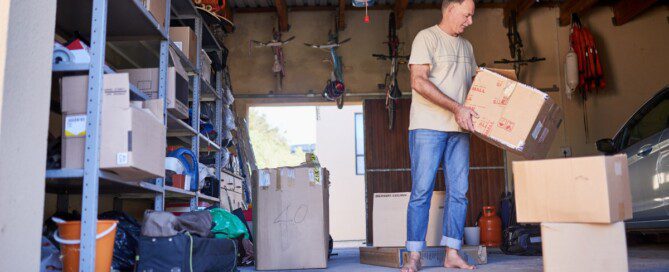 A man carries a cardboard box through a crowded garage filled with shelves, bicycles, luggage, and a parked car.