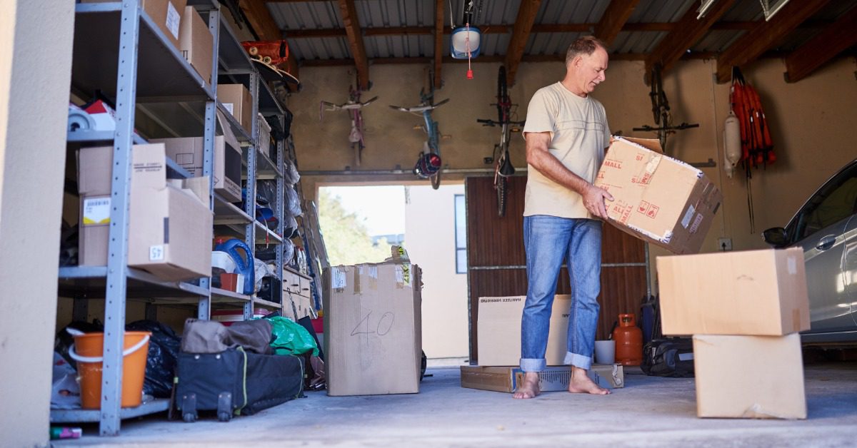 A man carries a cardboard box through a crowded garage filled with shelves, bicycles, luggage, and a parked car.