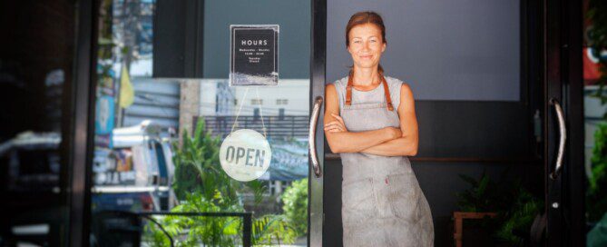 A female restaurant owner wearing an apron stands in an open cafe doorway beside a glass door with a sign.