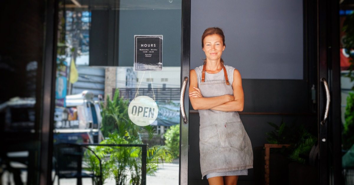 A female restaurant owner wearing an apron stands in an open cafe doorway beside a glass door with a sign.