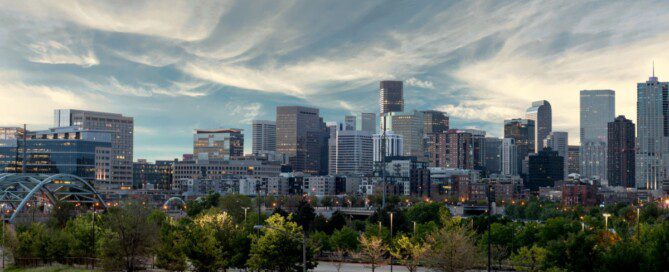 The Denver skyline rises beyond a green park as a curved bridge and roads frame the city beneath sweeping clouds.