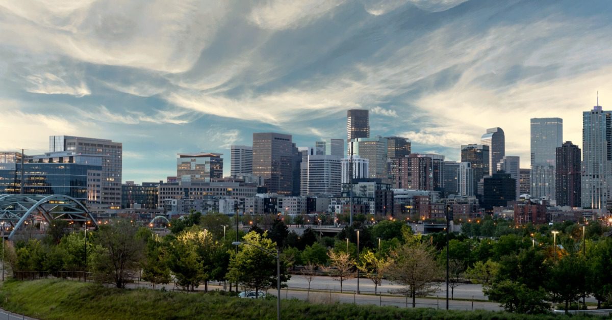 The Denver skyline rises beyond a green park as a curved bridge and roads frame the city beneath sweeping clouds.