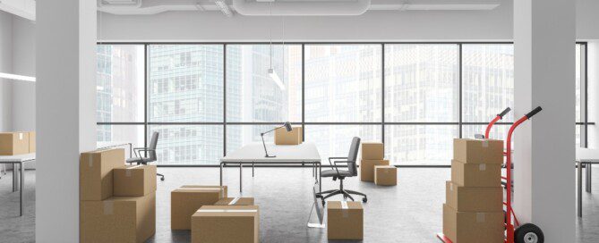 An office contains moving boxes, desks, chairs, and a loaded hand truck beside large windows overlooking city buildings.