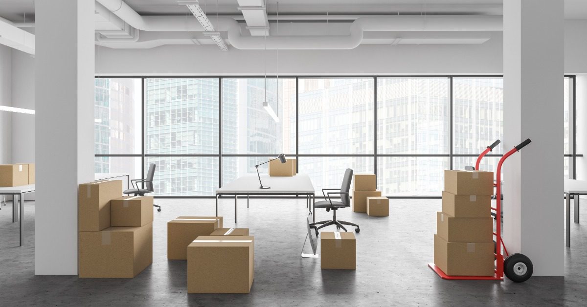 An office contains moving boxes, desks, chairs, and a loaded hand truck beside large windows overlooking city buildings.
