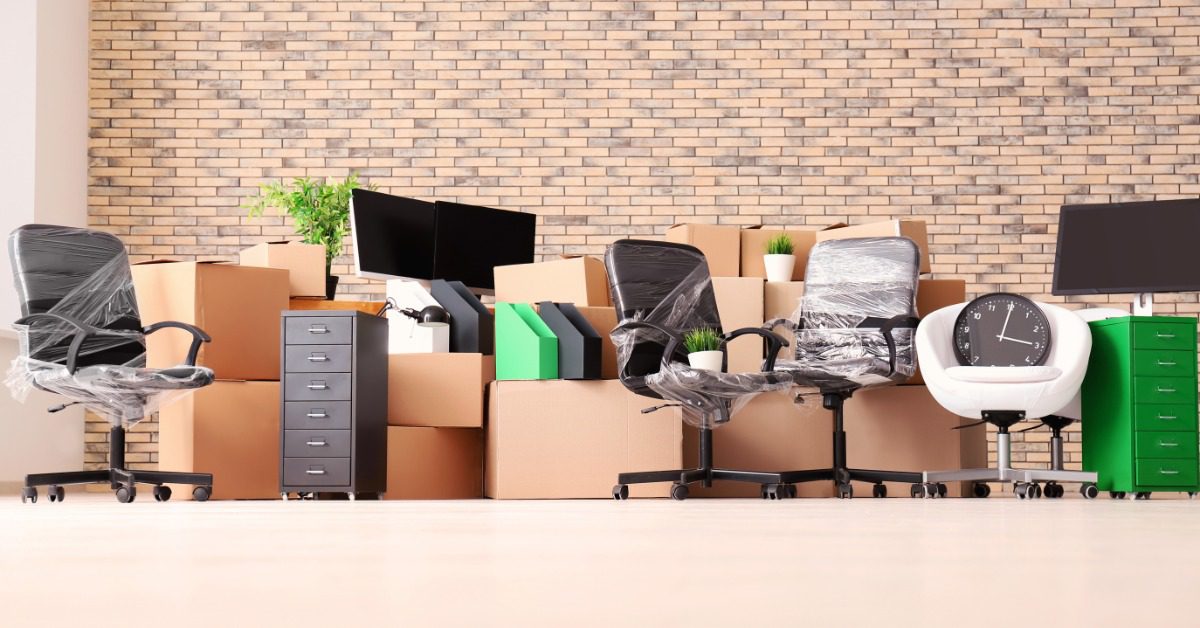 An office stores wrapped chairs, computer monitors, filing cabinets, and stacked boxes against a brick wall.