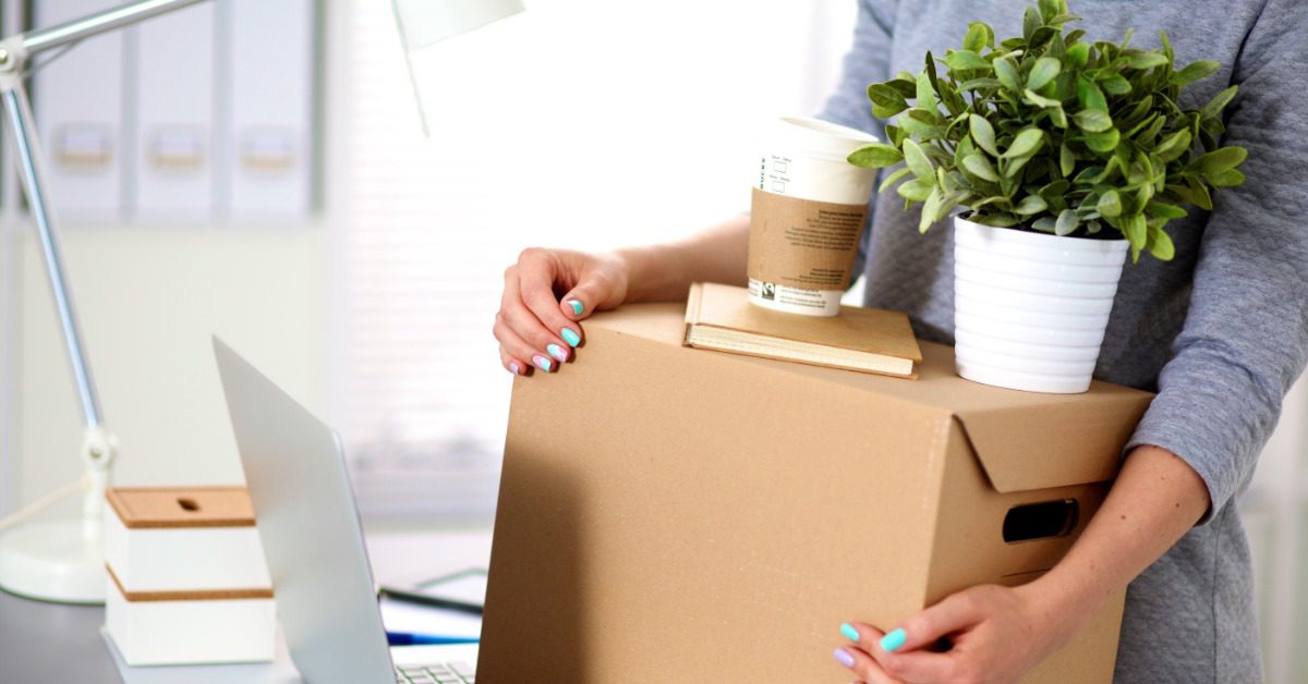 A woman carries a cardboard box with a plant and office items beside a laptop and filing cabinets in a bright workspace.