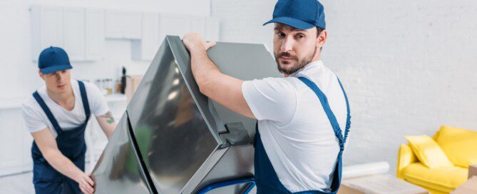 Two movers use a moving dolly to carry a large refrigerator through a bright living room during a move.