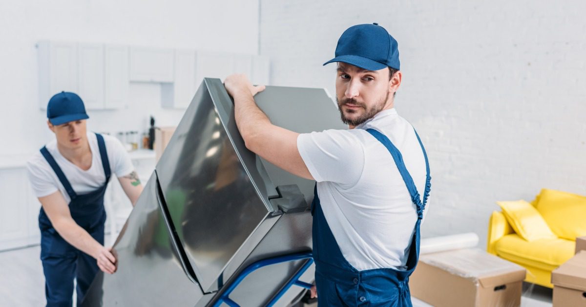 Two movers use a moving dolly to carry a large refrigerator through a bright living room during a move.