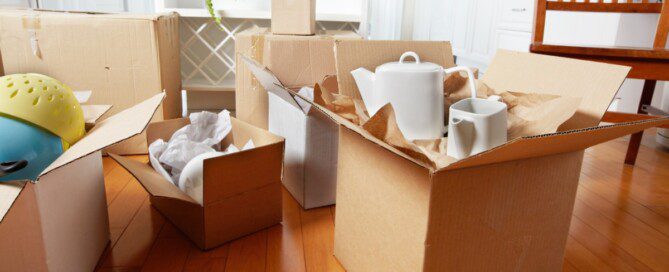 Open cardboard moving boxes filled with kitchen dishes and household items sit on a hardwood floor in a bright kitchen.