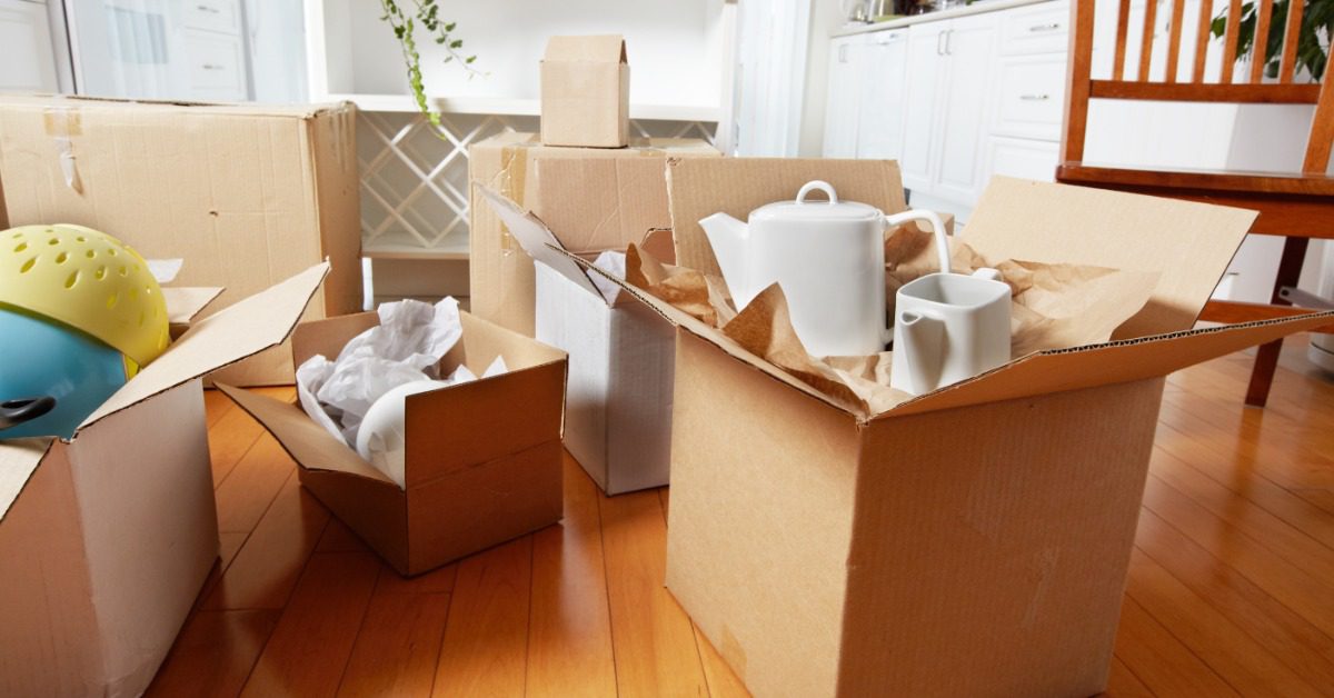 Open cardboard moving boxes filled with kitchen dishes and household items sit on a hardwood floor in a bright kitchen.