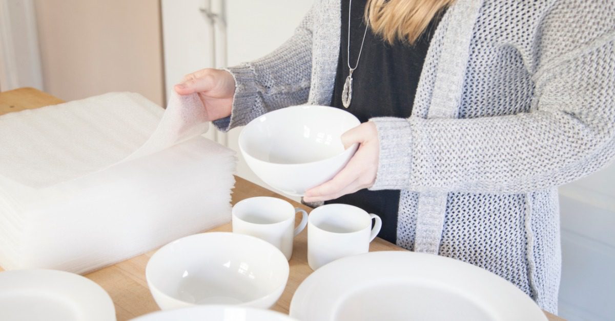 A woman carefully wraps a white ceramic bowl in protective packing material while preparing kitchen dishes for a move.