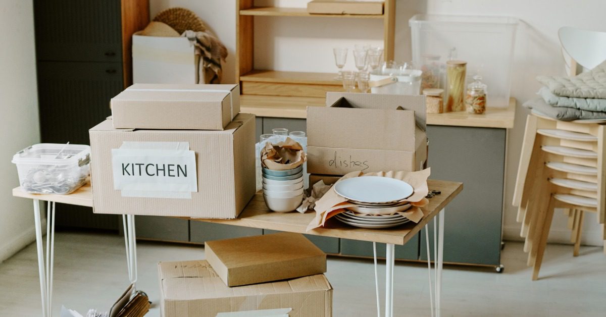 Cardboard moving boxes labeled "Kitchen" sit alongside stacked dishes and packing supplies in a partially packed room.