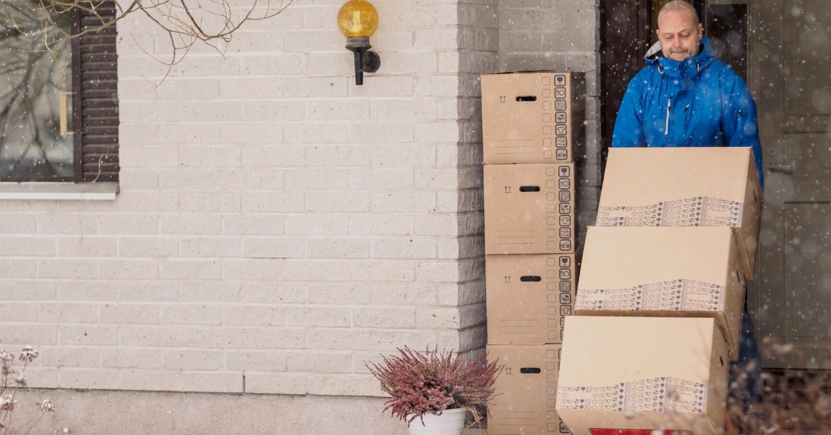 Man wearing a blue winter jacket moves stacked cardboard boxes on a hand truck outside a house during snowfall, with snow covering the ground.