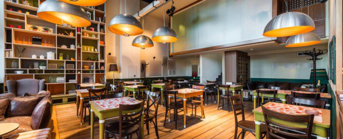 Modern restaurant dining room with wooden floors, colorful tables, large pendant lights, and floor-to-ceiling display shelving.