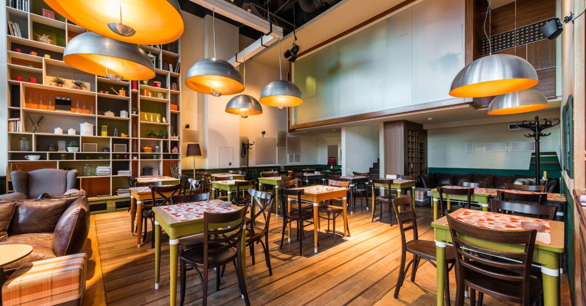 Modern restaurant dining room with wooden floors, colorful tables, large pendant lights, and floor-to-ceiling display shelving.