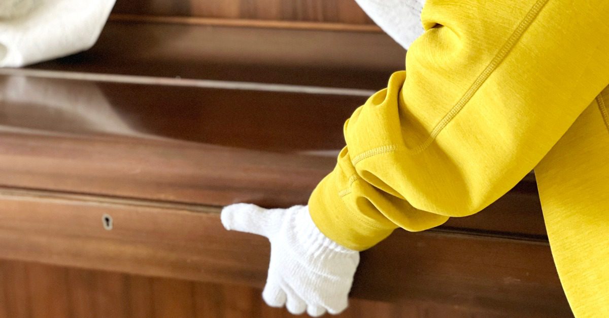 A gloved mover lifts a wooden piano while another their other hand pulls a breathable padded sheet over the piano.