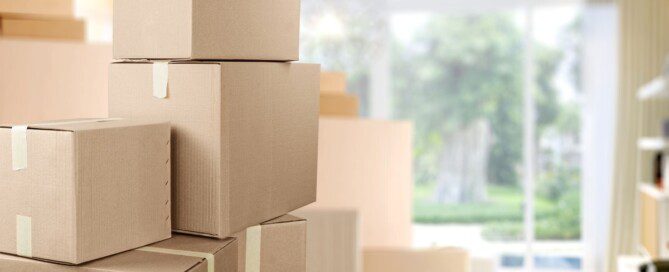 A stack of taped cardboard moving boxes fills a bright room as sunlight shines through a large window.